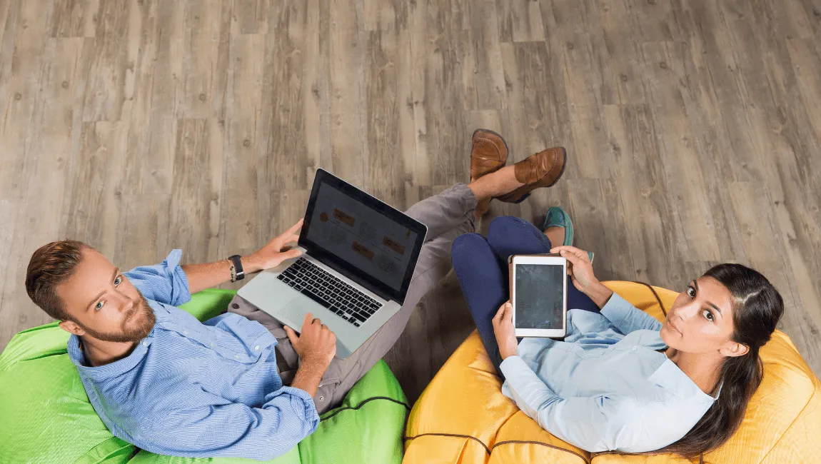 A man and woman sit on colorful bean bags, each using a laptop in a relaxed indoor setting.