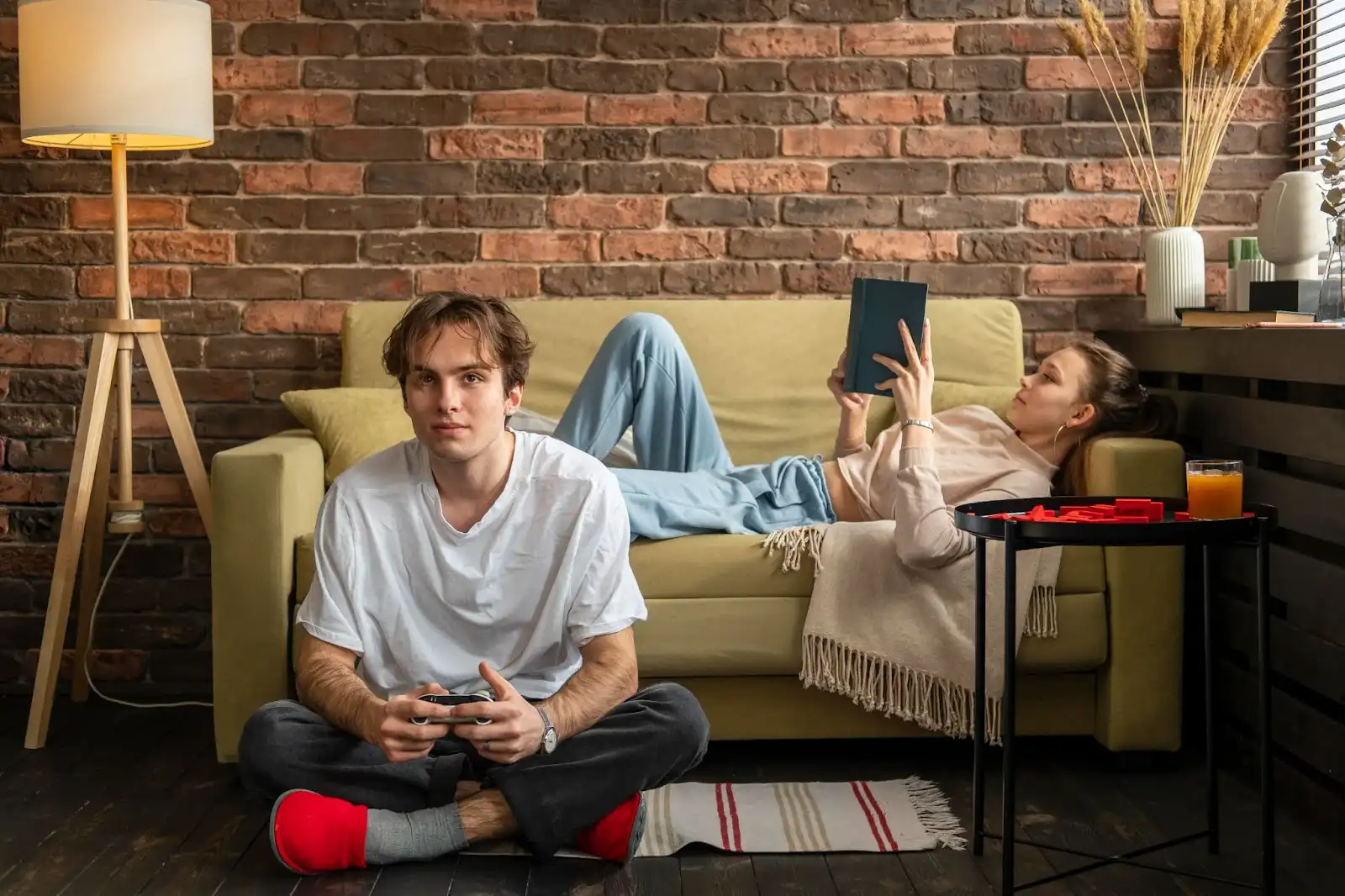 Young woman browsing the internet on a tablet while relaxing on a sofa, with a man in the front playing a video game — illustrating modern home internet usage.
